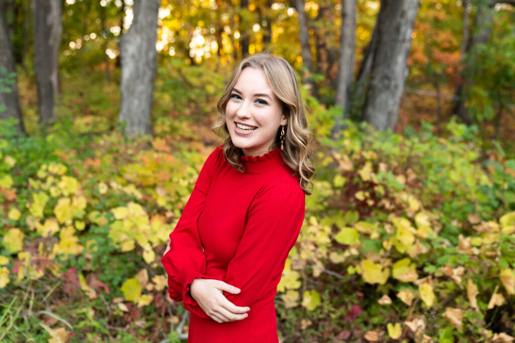 Teen girl wearing a red dress laughs at the camera during her senior photo shoot. She's surrounded by fall foliage. This was taken at the Minnesota Landscape Arboretum in Chaska, Minnesota