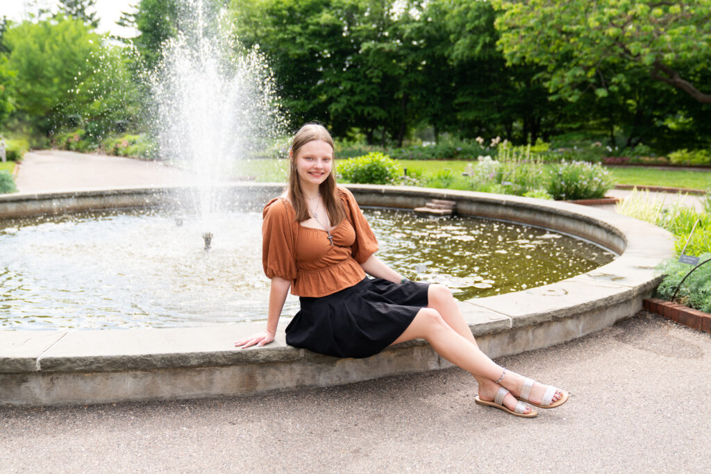 teen girl poses next to a fountain for her senior pictures at the Minnesota Landscape Arboretum in Chaska, Minnesota.