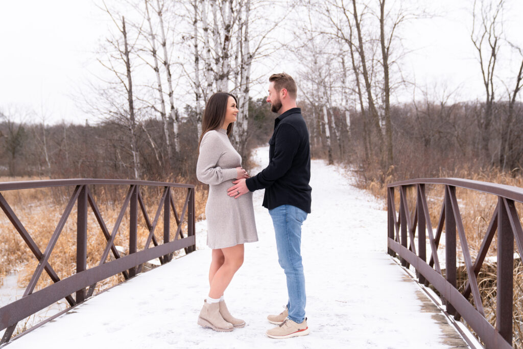A couple standing on a bridge looks at each other while holding onto the woman's baby bump during their winter maternity session.