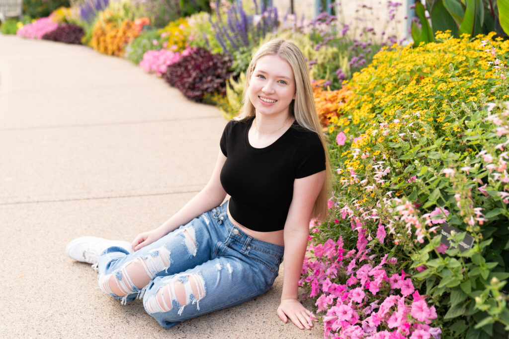 Teen girl sits on the sidewalk next to a beautiful flower bed during her senior photos.