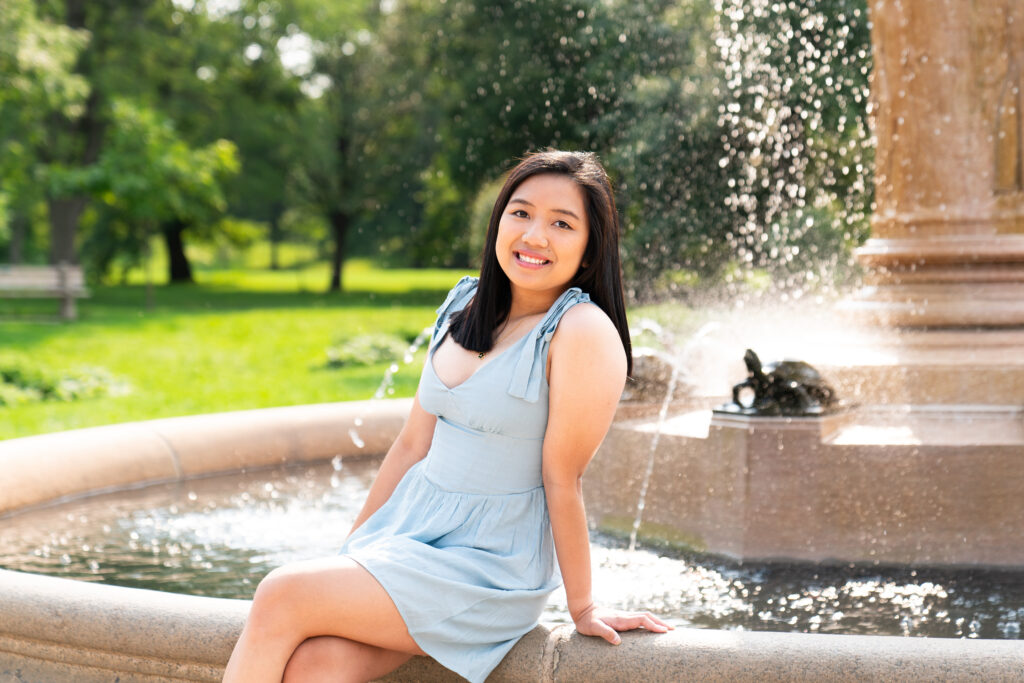 A teen girl sits on a fountain and smiles at the camera during her senior pictures at the Lyndale Rose Gardens in Minneapolis, Minnesota.