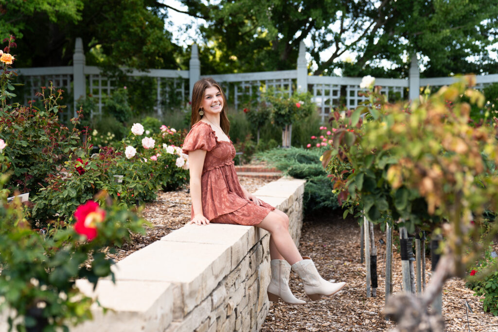 A high school aged girl sits on a ledge in a rose garden wearing a rust colored dress as one of her senior picture outfits.