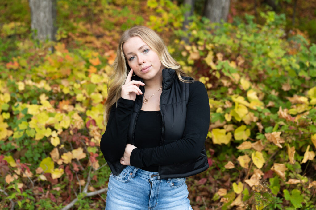 A high school girl wearing a black top, a black vest, and jeans poses among fall colors during her senior photos.