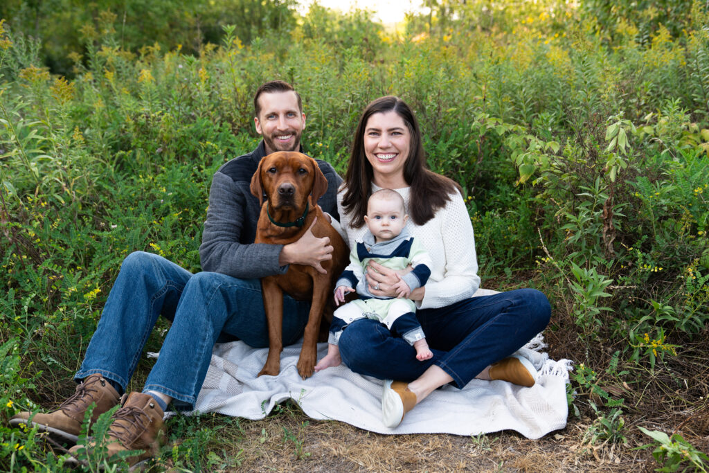 A family sits together on a blanket surrounded by tall grasses for their fall family photo shoot.