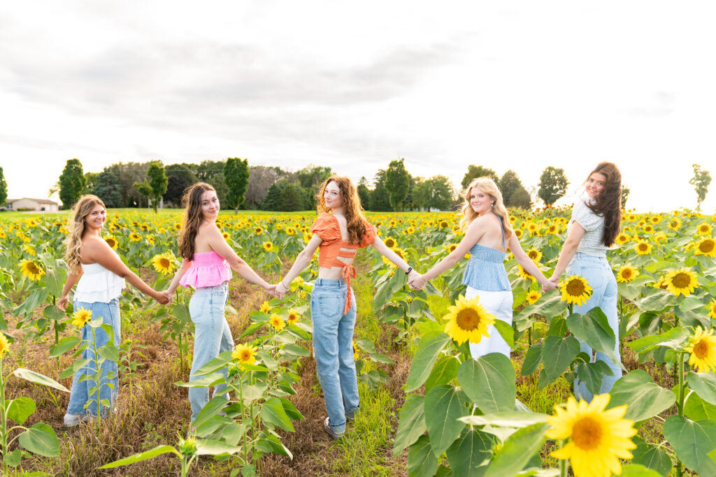 Five teen girls hold hands in a sunflower field and look back towards the camera during their senior rep photo shoot.